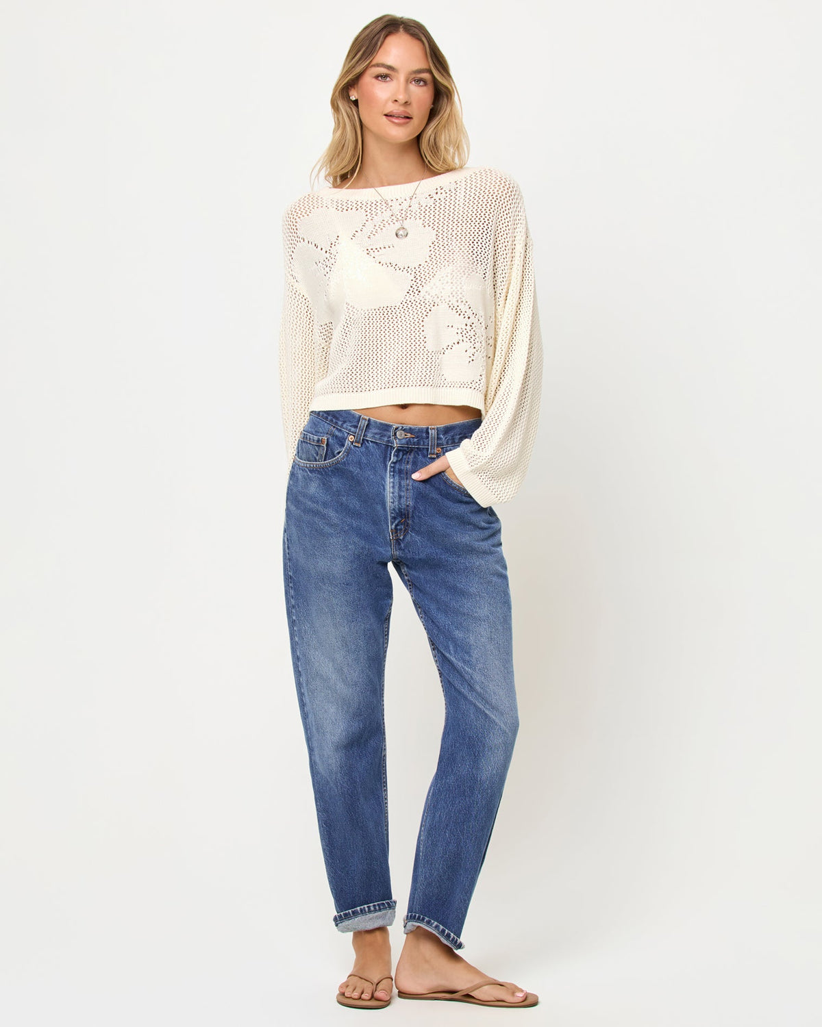Front view of a model wearing the LSPACE Island Time Sweater in Cream, styled with jeans, featuring an open-knit crochet design with a large floral motif and long sleeves.